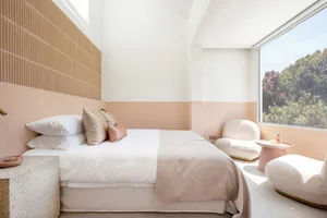 A modern bedroom featuring a bed with white bedding, white-cement walls, and a view of trees through a window.