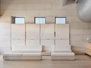 Minimalist living room with white sofas and wooden bench seating, glass windows, and a modern light fixture.