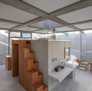 A minimalist living room with concrete walls, wooden stairs, and a glass partition.
