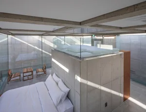 A minimalist bedroom featuring a platform bed with a glass railing, surrounded by concrete walls and floors.