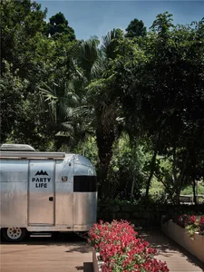 A silver, metallic camping trailer with the text 'PARTY LIFE' on its side, parked on a wooden deck surrounded by lush greener