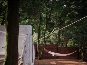 A white hammock hanging from a wooden frame on a deck surrounded by trees and foliage.