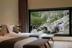 A well-lit hotel room with a view of greenery and rocks outside through a large window.