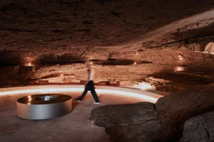A person is walking past a circular metal table in a cave-like interior space with rock formations and illuminated seating.