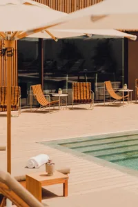 A minimalist poolside lounge area with wooden lounge chairs, glass tables, and umbrella shades.