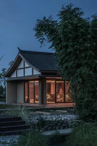 A minimalist residential home with a traditional roof and a warm, inviting interior, illuminated at night, surrounded by lush