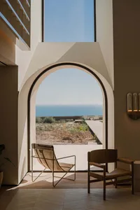 A minimalist living room with a concrete floor and a large arched window overlooking the sea.