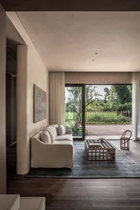 A modern living room with a beige sofa, a wooden coffee table, and a large window overlooking a green outdoor area.