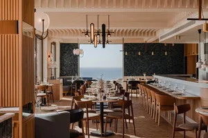 Modern restaurant interior with sea view, featuring wooden tables, seating, and minimalist decor.
