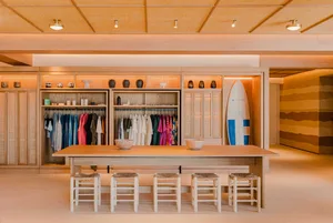 A retail space featuring wooden shelves, stools, and a table, illuminated by warm lighting, with a surfboard leaning against 
