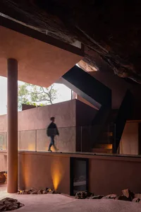 A modern living space featuring concrete walls and flooring, with a person walking on a glass-walled balcony.