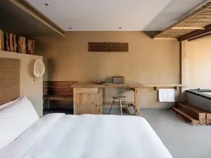 A minimalist bedroom featuring wooden furniture, a large bed with a white bedspread, and a table with a wooden stool and a sp