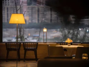 Interior of a cozy restaurant with a view through a large window, featuring wooden chairs, glass tables, and fabric seating, 