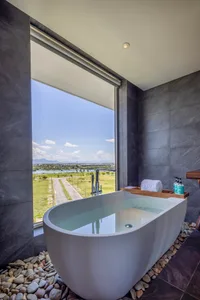 A modern bathroom with a white freestanding bathtub surrounded by rocks, featuring a large window overlooking a peaceful land