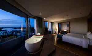 A modern bedroom with a white bed, wooden floor, and large windows overlooking a cityscape at dusk.