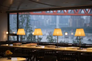 A well-lit cafe interior with wooden tables and chairs, warm lighting, and a view of a bridge over a body of water.