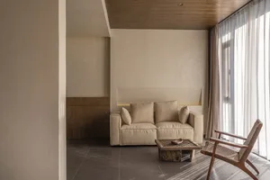 A minimalist living room with a brown sofa, beige cushions, and a wooden coffee table in front of a window with white curtain