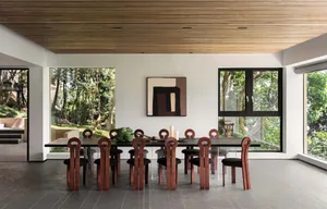 Modern dining room featuring wooden chairs and a glass dining table surrounded by large windows and greenery.