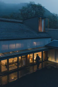A modern house with a glass exterior at night with lights on and a person walking outside.