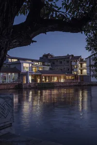 A residential house overlooking a river with glass windows reflecting the interior lights and the surrounding trees.