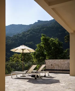 A photo of an outdoor space with stone cladding, two lounge chairs, and an umbrella, overlooking a lush, forested mountain.