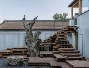 A natural wooden staircase with a large, textured tree trunk in the center, surrounded by rocks, and leading up to a modern b