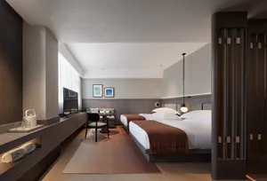 A minimalist bedroom with a bed covered in a brown throw blanket, a small table with a vase, and a chair in front of it.