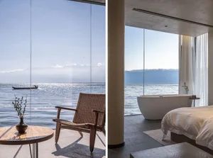 A minimalist residential interior with a concrete floor, a freestanding bathtub overlooking the water, a bed with white linen