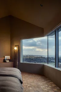 A modern bedroom featuring a wooden floor, a bed with a gray blanket, and a window overlooking a rocky coastline.