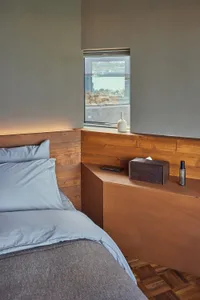 Modern minimalist bedroom featuring a wooden headboard and grey bedding, with a small window overlooking a water view.