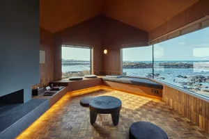 A modern, wooden-floored room with a view of the sea, featuring seats and a circular table.