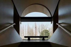 Concrete staircase viewed from above, featuring an arched window with a bench and a scenic outdoor view of bare trees.