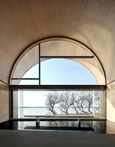 Modern minimalist interior space featuring glass and concrete materials with a view of a serene water body and leafless trees