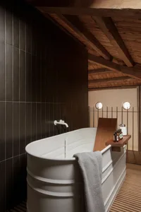 A modern minimalist bathroom with a white concrete bathtub, dark tiled walls, and wooden beams on the ceiling.