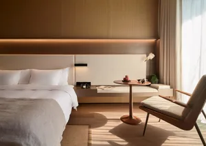 A minimalist bedroom featuring a bed with wooden headboard, white bedding, and a wooden side table with a book and coffee cup