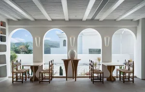 A minimalist restaurant interior with wooden tables and chairs, open windows, and a view of the mountains.