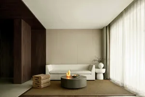 A modern living room with a minimalist sofa, round coffee table, and cushioned floor seating. Large windows with white curtai