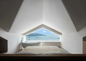 A modern minimalist bedroom with concrete walls featuring a large window overlooking the ocean.