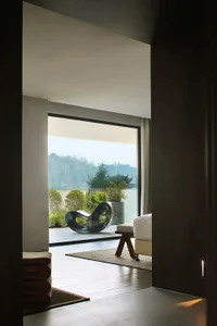 A modern minimalist bedroom with wood flooring, a white bed, and a black sculptural chair on a balcony overlooking a scenic l