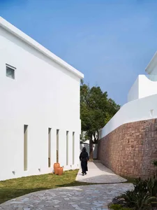 Person walking on a paved pathway alongside a minimalist residential building with white plaster walls and stone walls.