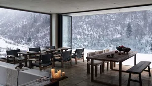 A cozy winter lodge dining room featuring wooden tables, black leather chairs, candles, and a view of snowy mountains.