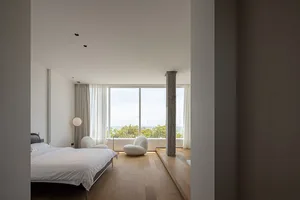 A modern bedroom with a white bed, white chairs, and wooden flooring, featuring a large window with curtains and a concrete c
