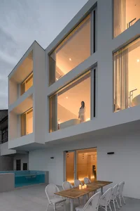 A modern residential house with concrete and glass structures, featuring a large window and outdoor seating area.