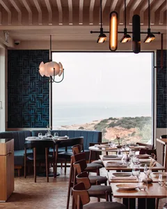 A modern restaurant dining room with sea views, wooden tables, fabric seating, and glass chandeliers.