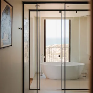 Minimalist bathroom featuring a white tiled floor and walls, with a large oval-shaped bathtub overlooking the ocean.
