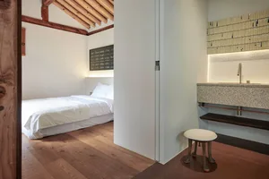 Minimalist bedroom with white walls, light wooden floor, white bedding, and a wooden stool under a granite countertop with a