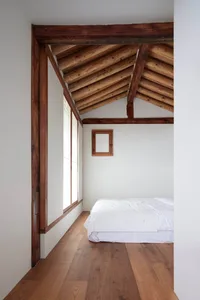 A minimalist bedroom with white walls, wooden floors, and a single white bed against a white wall. Wooden beams in the ceilin