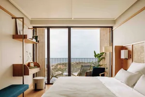 A serene minimalist bedroom with a large window overlooking the sea and a wooden chair with a green cushion.