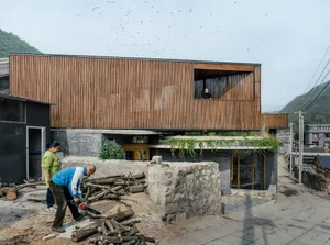 People cutting logs with a chainsaw in front of a modern building with wooden exterior and concrete walls.