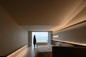 A minimalist bedroom with soft lighting, featuring a large window overlooking the ocean.